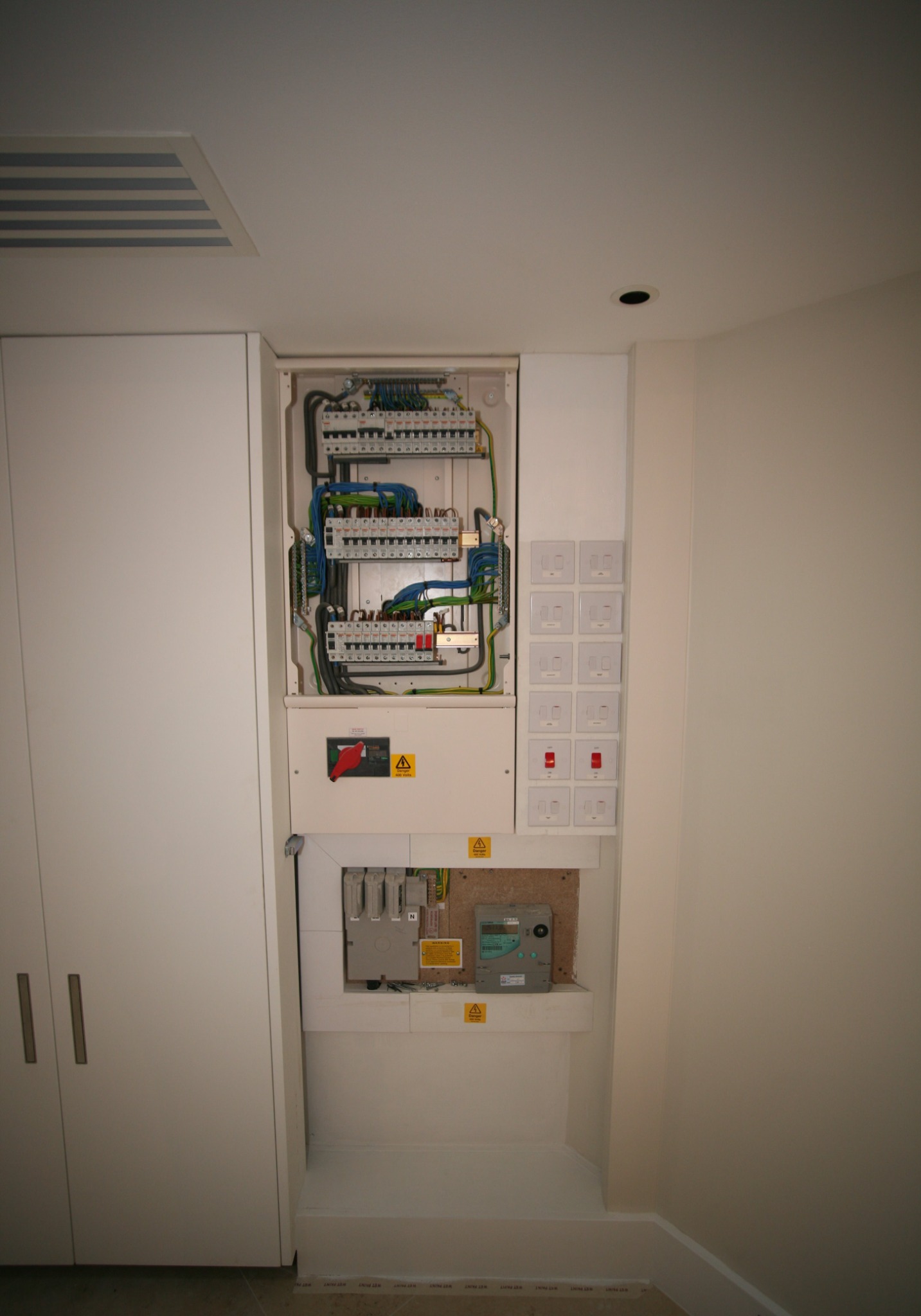 Otwarty panel elektryczny z widocznymi bezpiecznikami, przewodami i licznikiem, zamontowany w ścianie obok białej szafy w pomieszczeniu o jasnych ścianach.