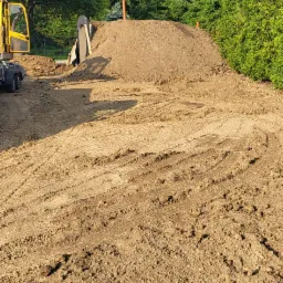Żółta koparka częściowo widoczna obok hałdy piasku z dwiema pionowymi rurami w Jaśle, teren przygotowywany pod budowę.