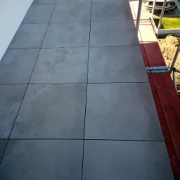 Szare płytki elewacyjne ułożone na balkonie, widok z góry, fragment rusztowania i samochód w tle.