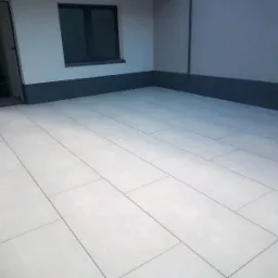 Wykończona posadzka z jasnych, prostokątnych płytek na zewnątrz budynku. Widoczne okno i drzwi wejściowe. Minimalistyczny styl wykończenia.