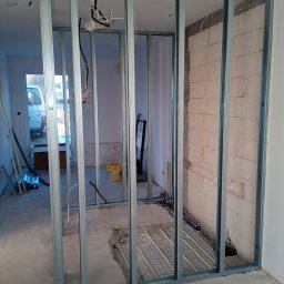 ,,Cztery Kąty'' Patryk Piątek - Metalowa konstrukcja ścianki działowej w surowym wnętrzu, widoczne przewody elektryczne i instalacja ogrzewania podłogowego. Ściana z bloczków.
