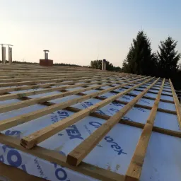 Konstrukcja drewniana dachu w trakcie budowy z widoczną membraną dachową i kominami w tle, sfotografowana w słoneczny dzień.