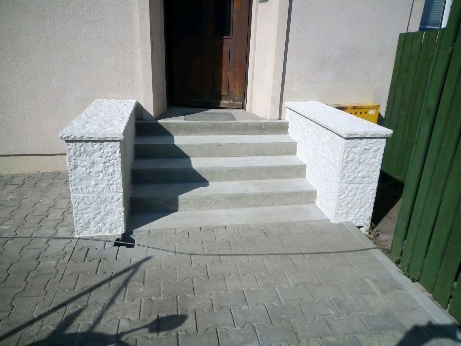 Betonowe schody wejściowe z dekoracyjnymi, tynkowanymi balustradami, prowadzące do ciemnych drzwi wejściowych budynku, z widoczną kostką brukową przed schodami i zielonym płotem w tle.