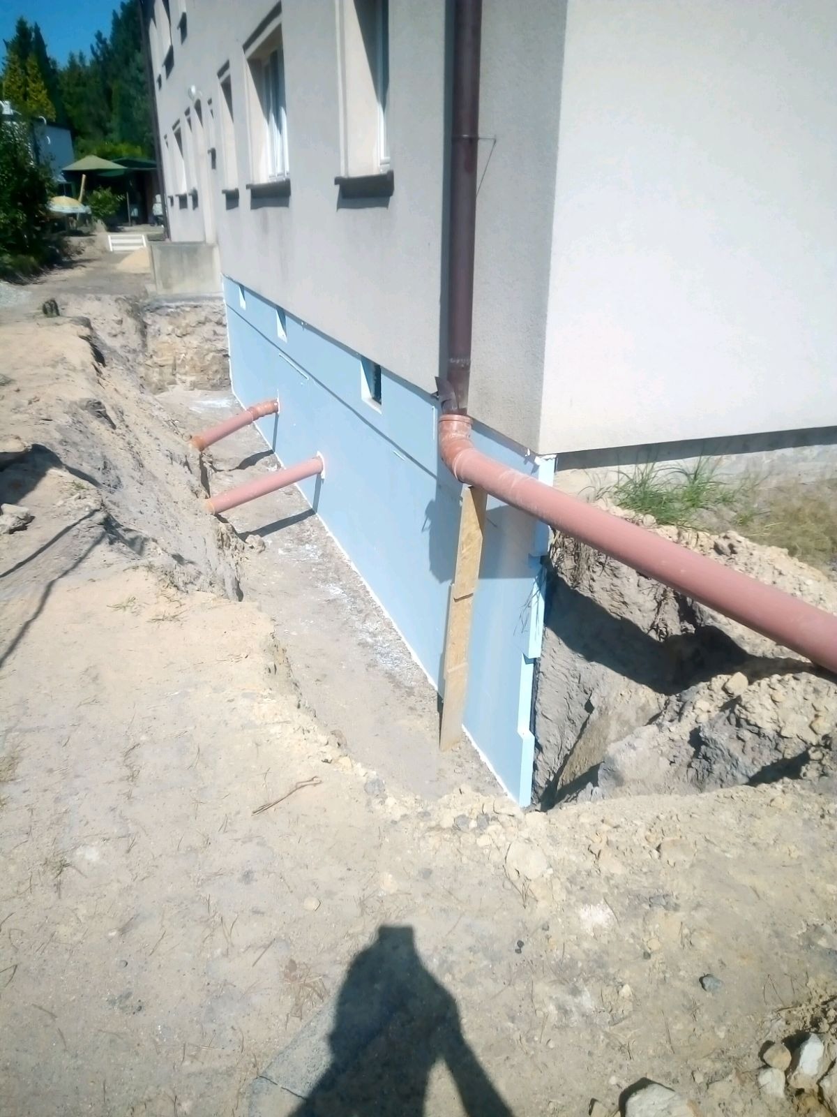 Fundament budynku ocieplony niebieskimi płytami izolacyjnymi, widoczne rury drenażowe wychodzące ze ściany, wykop wzdłuż fundamentu, cień osoby fotografującej na pierwszym planie.