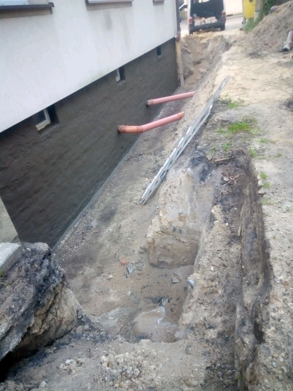Fundament budynku po izolacji, z widocznymi rurami drenażowymi i wykopem w ziemi, drabina oparta o ścianę.