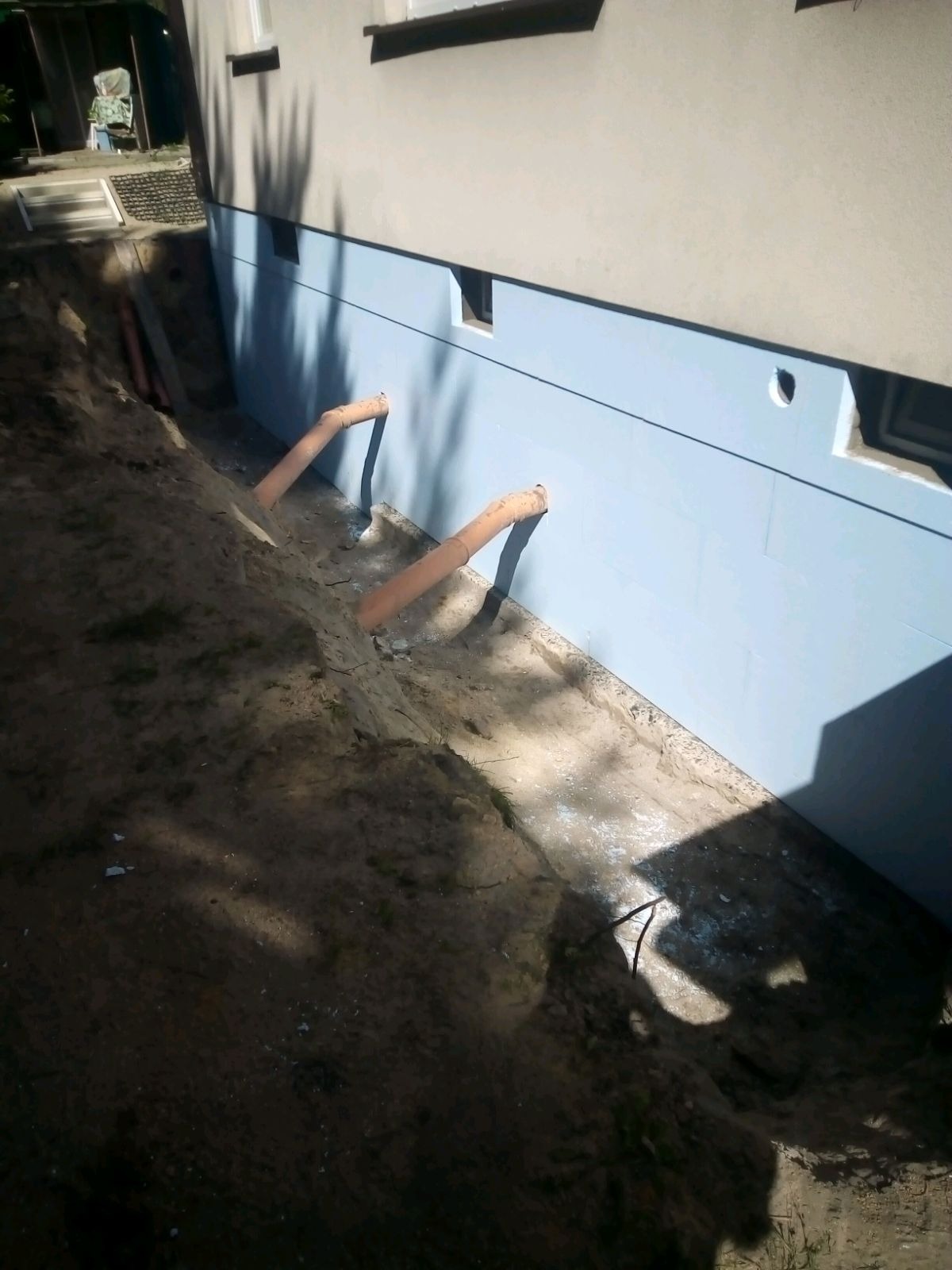 Odsłonięty fundament budynku z niebieską izolacją, wystające rury drenażowe, wykop w ziemi.