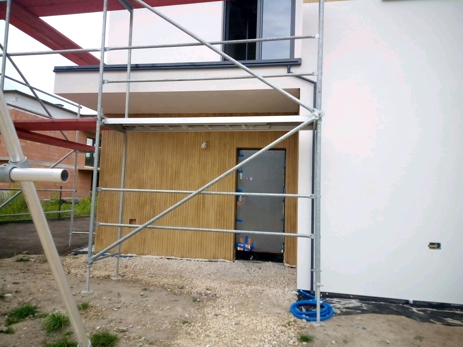 Budynek w trakcie prac elewacyjnych, widoczne rusztowanie, fragment otynkowanej ściany na biało, drewniana okładzina elewacyjna i stalowe drzwi wejściowe zabezpieczone folią.