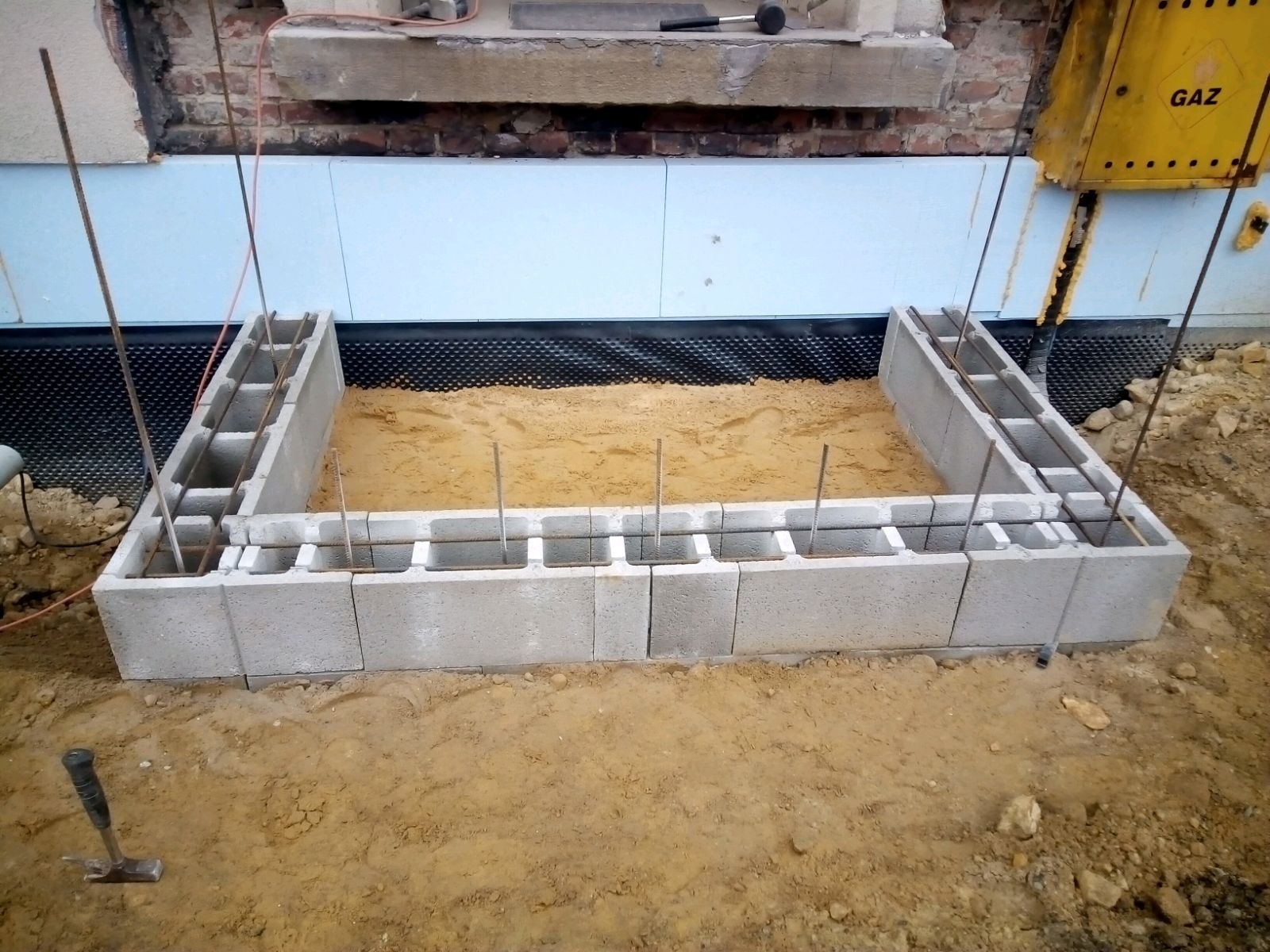 Fundament z bloczków betonowych zbrojony stalą, piasek wewnątrz, zabezpieczony folią kubełkową, widoczna instalacja gazowa oraz narzędzie budowlane na ziemi.