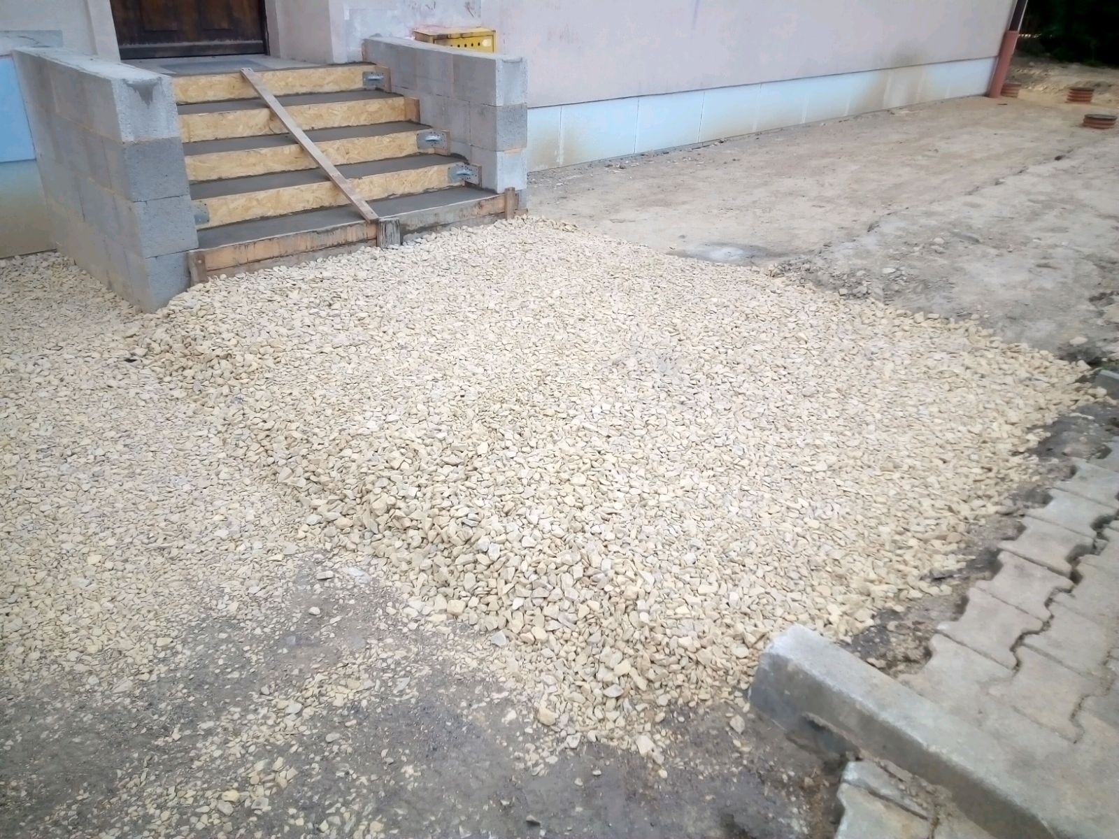 Betonowe schody wejściowe w trakcie budowy, z szalunkiem i częściowo wypełnione betonem, otoczone jasnym żwirem.