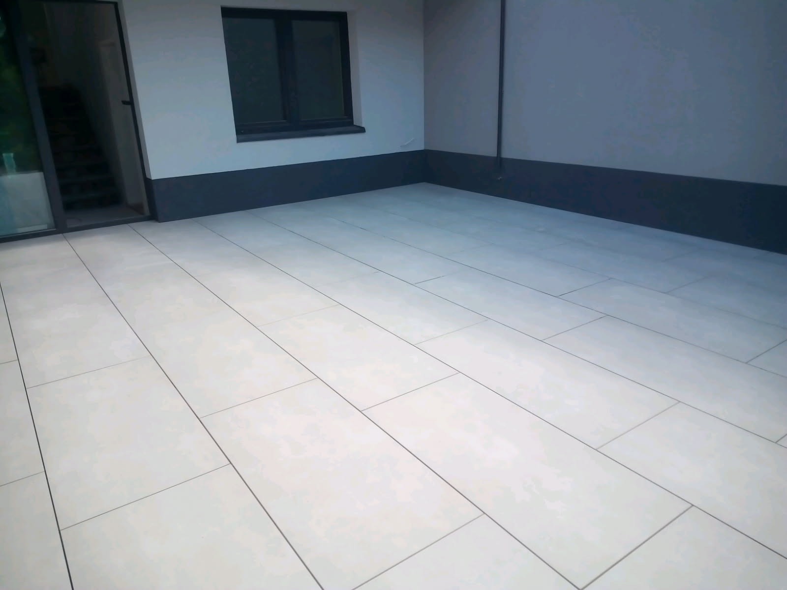 Wykończona posadzka z jasnych, prostokątnych płytek na zewnątrz budynku. Widoczne okno i drzwi wejściowe. Minimalistyczny styl wykończenia.