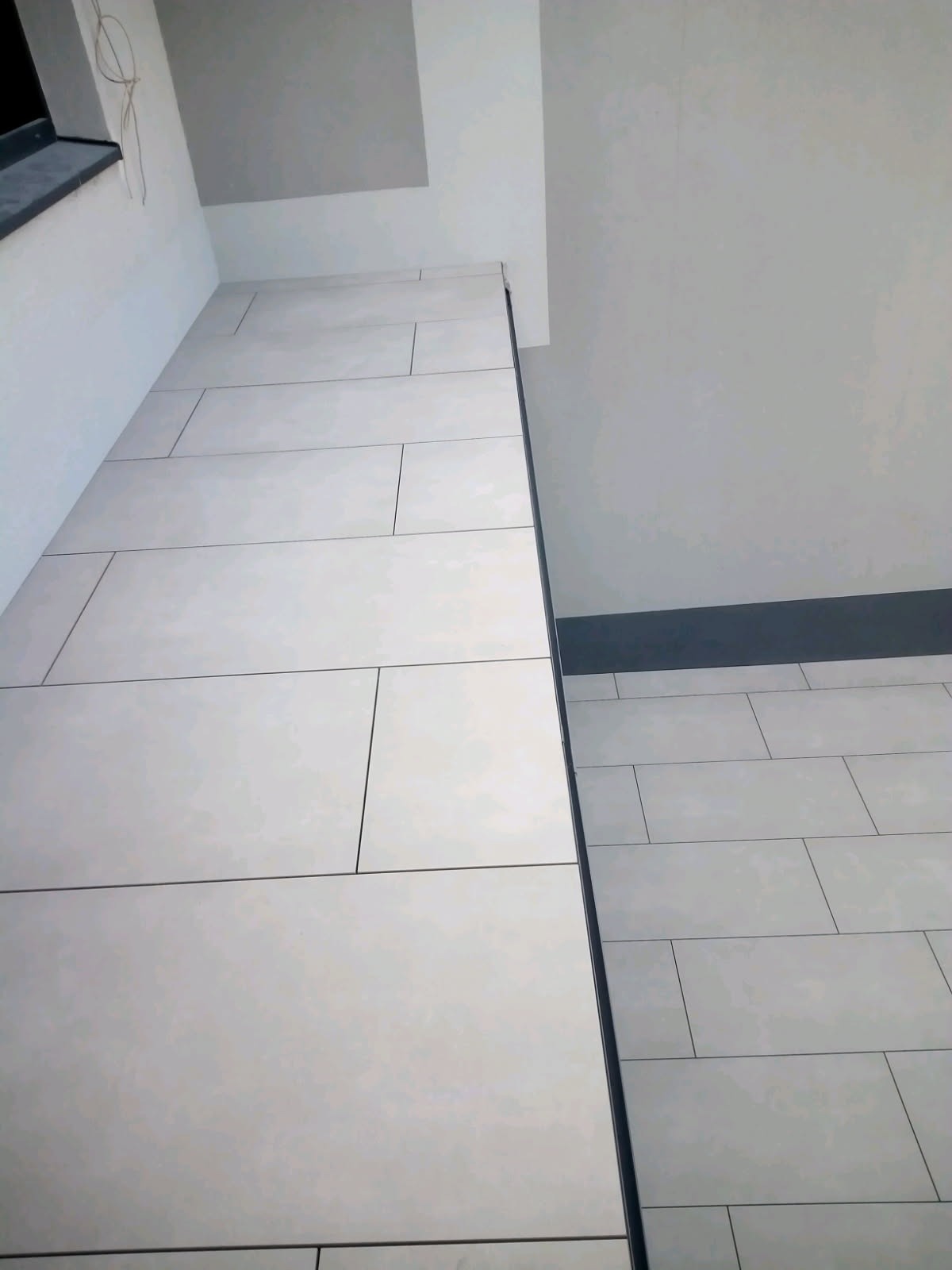 Ściana i podłoga wyłożone jasnymi, prostokątnymi płytkami. Minimalistyczny design z widocznymi fugami. Ujęcie pod kątem, ukazujące połączenie powierzchni.