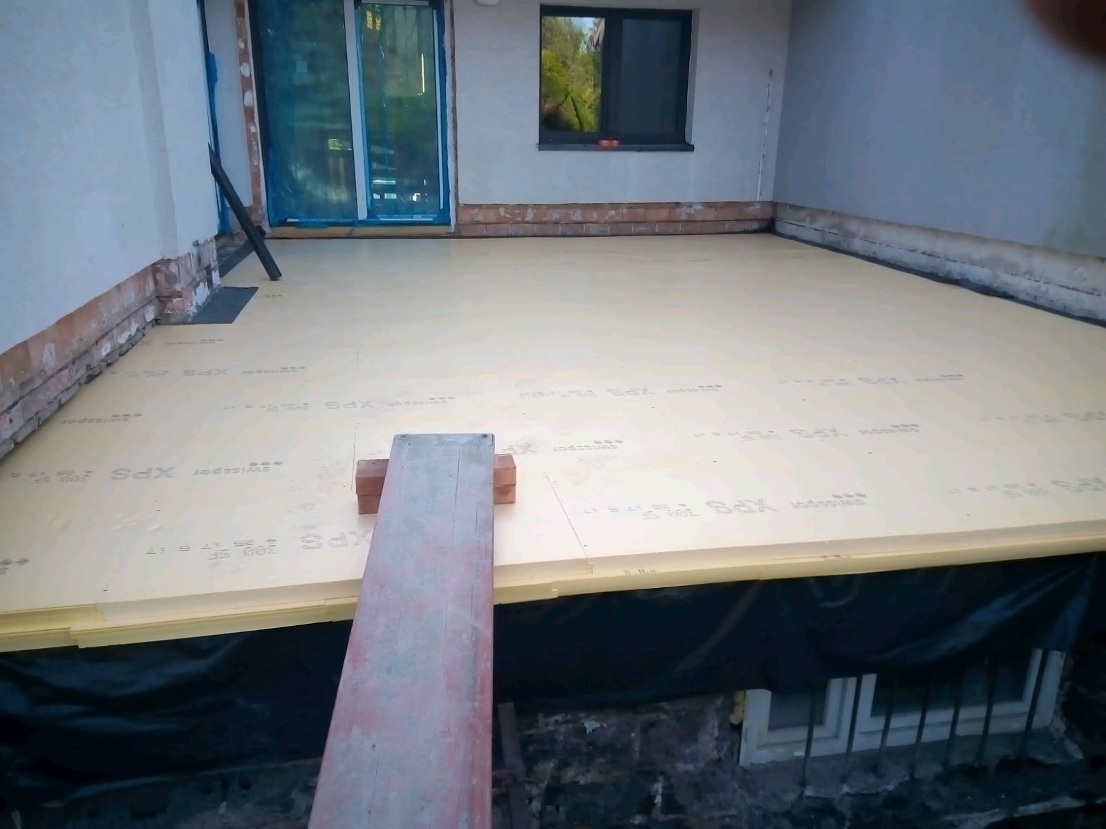 Izolacja termiczna podłoża z płyt XPS Swissopor, widoczne okno piwniczne, ściany z ceglaną podmurówką i deska robocza. Etap prac przed wylewką.