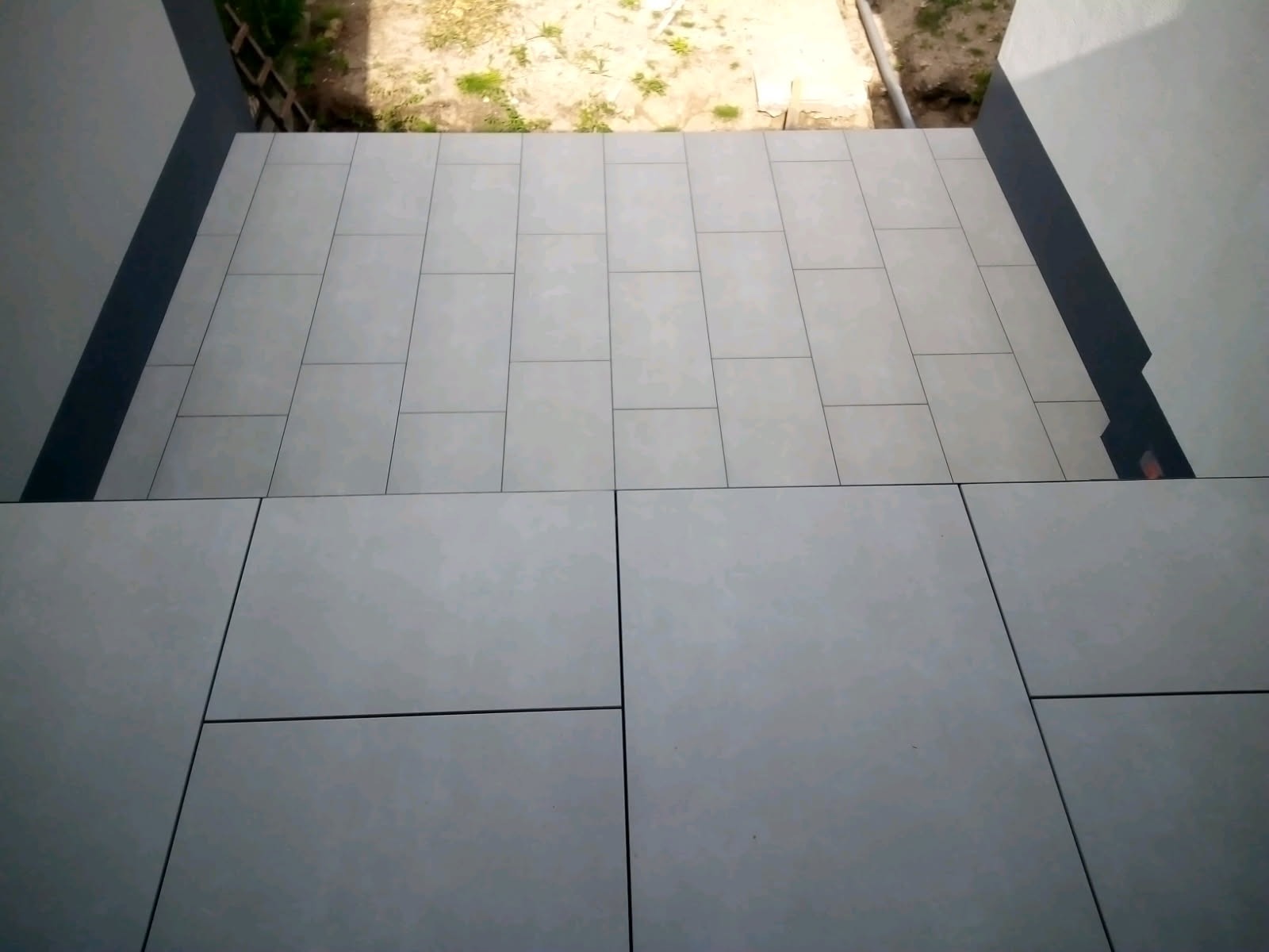 Wykończenie balkonu: jasne, prostokątne płytki ułożone na posadzce balkonu, widok z góry, z fragmentem elewacji budynku po bokach. Minimalistyczny design.