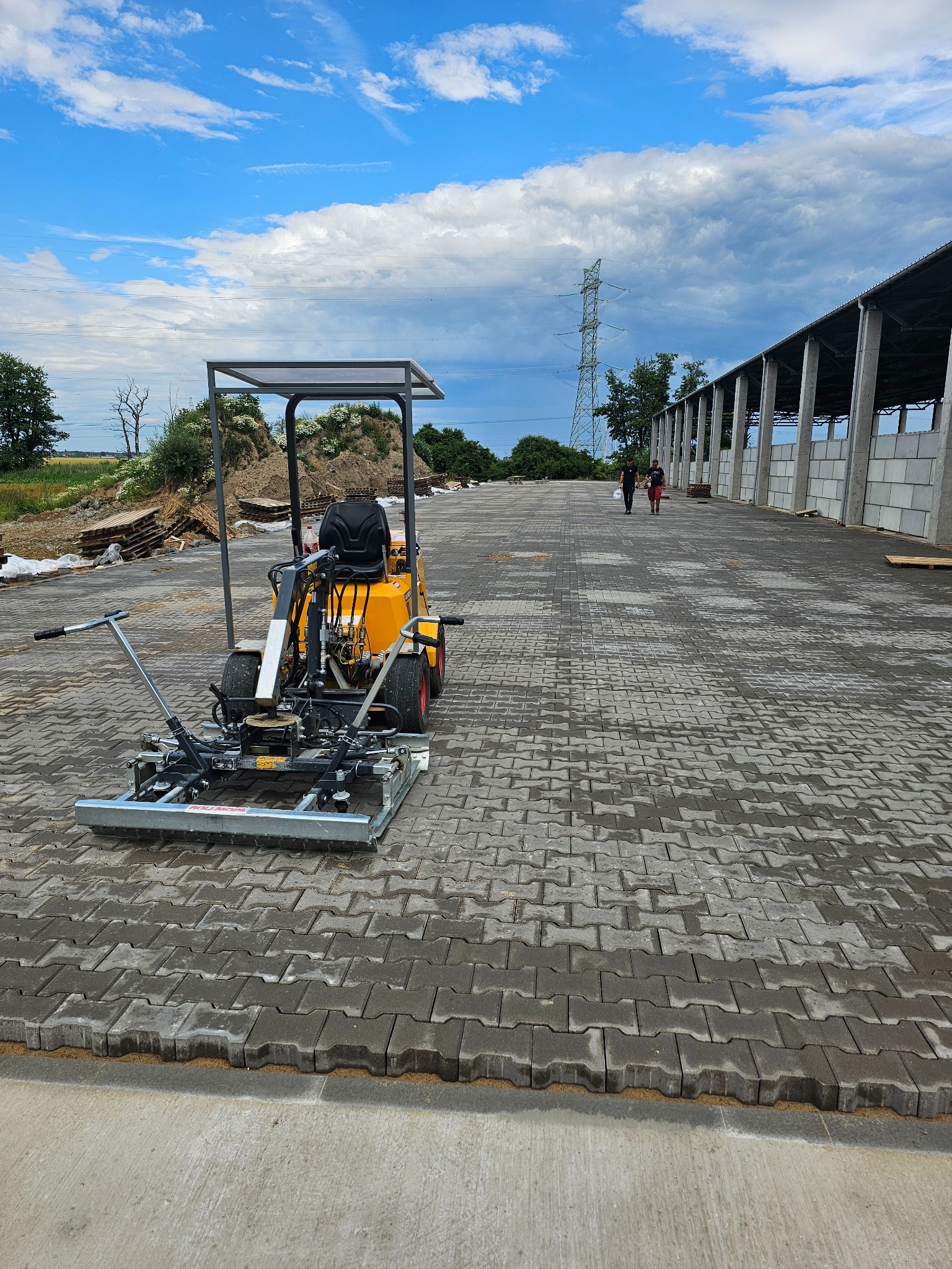Żółta maszyna do układania kostki brukowej na tle nowo ułożonej, szarej kostki w kształcie litery H, widoczni dwaj mężczyźni w oddali, słupy wysokiego napięcia i budynek gospodarczy.