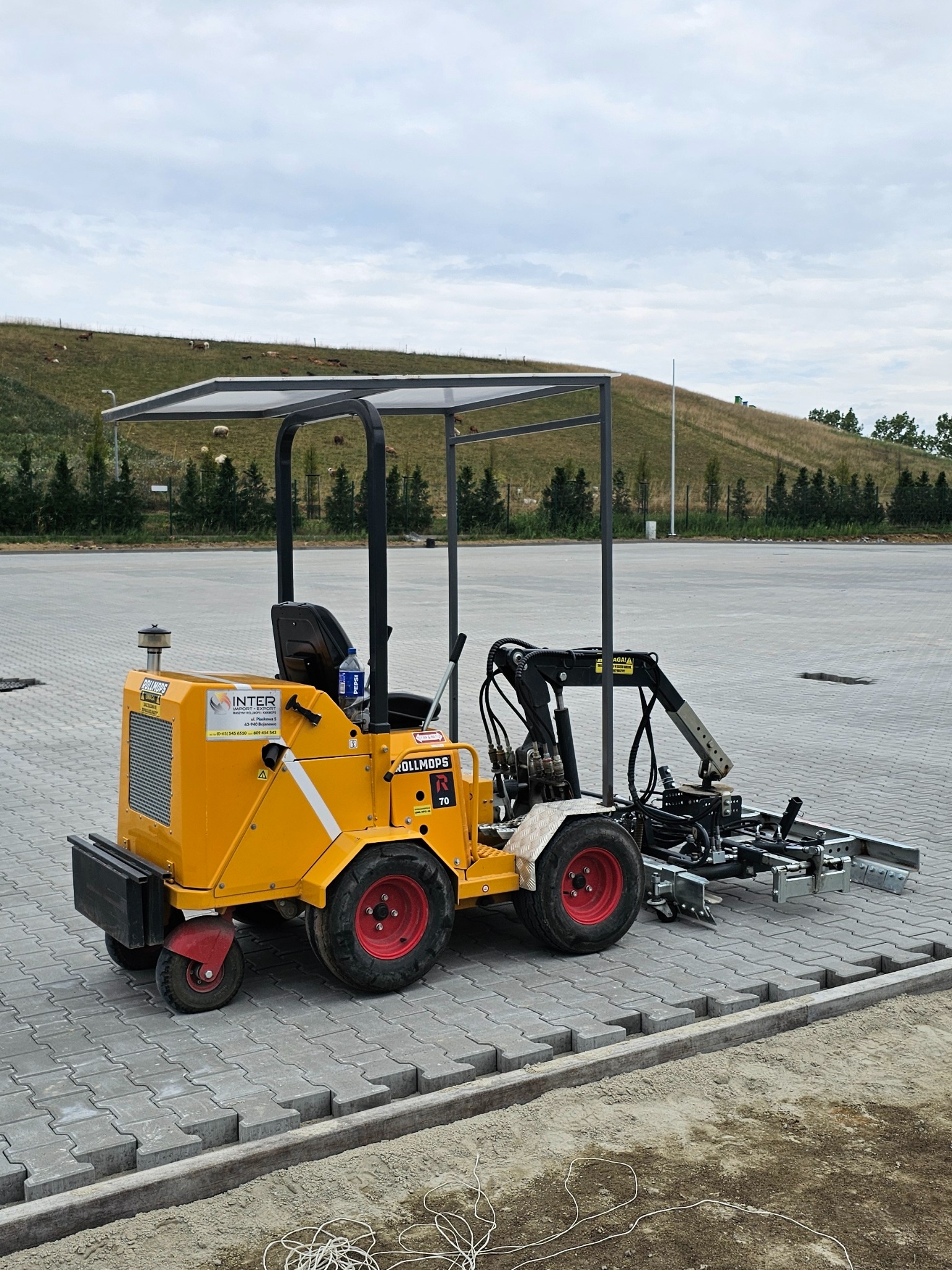 Żółta maszyna do układania kostki brukowej Rollmops R70 z czerwonymi kołami na placu, w tle zielone wzgórze z pasącymi się owcami.