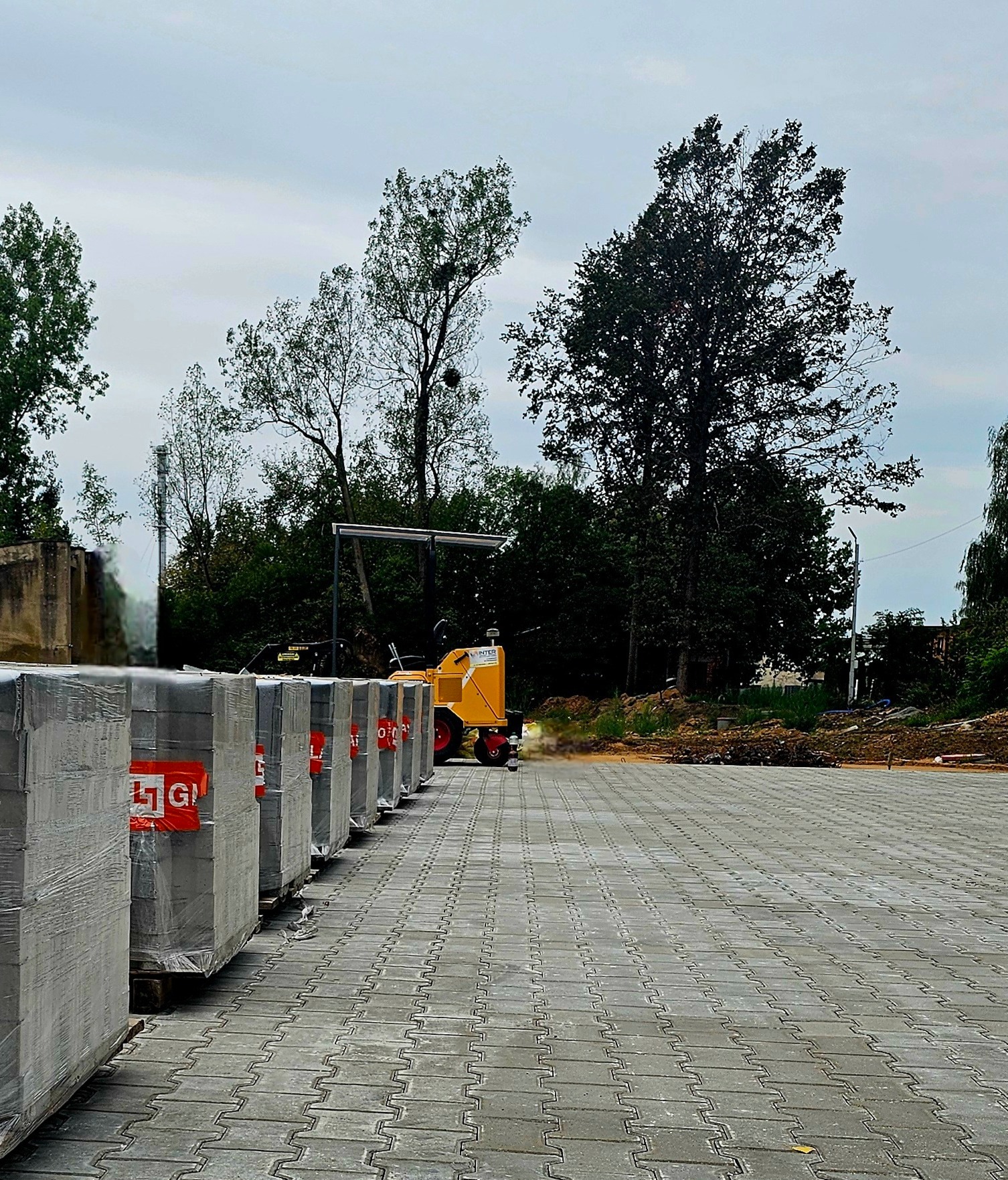 Ułożona szara kostka brukowa typu puzzle, palety z zapakowaną kostką z logo producenta w tle, oraz maszyna do układania kostki brukowej na świeżym powietrzu.