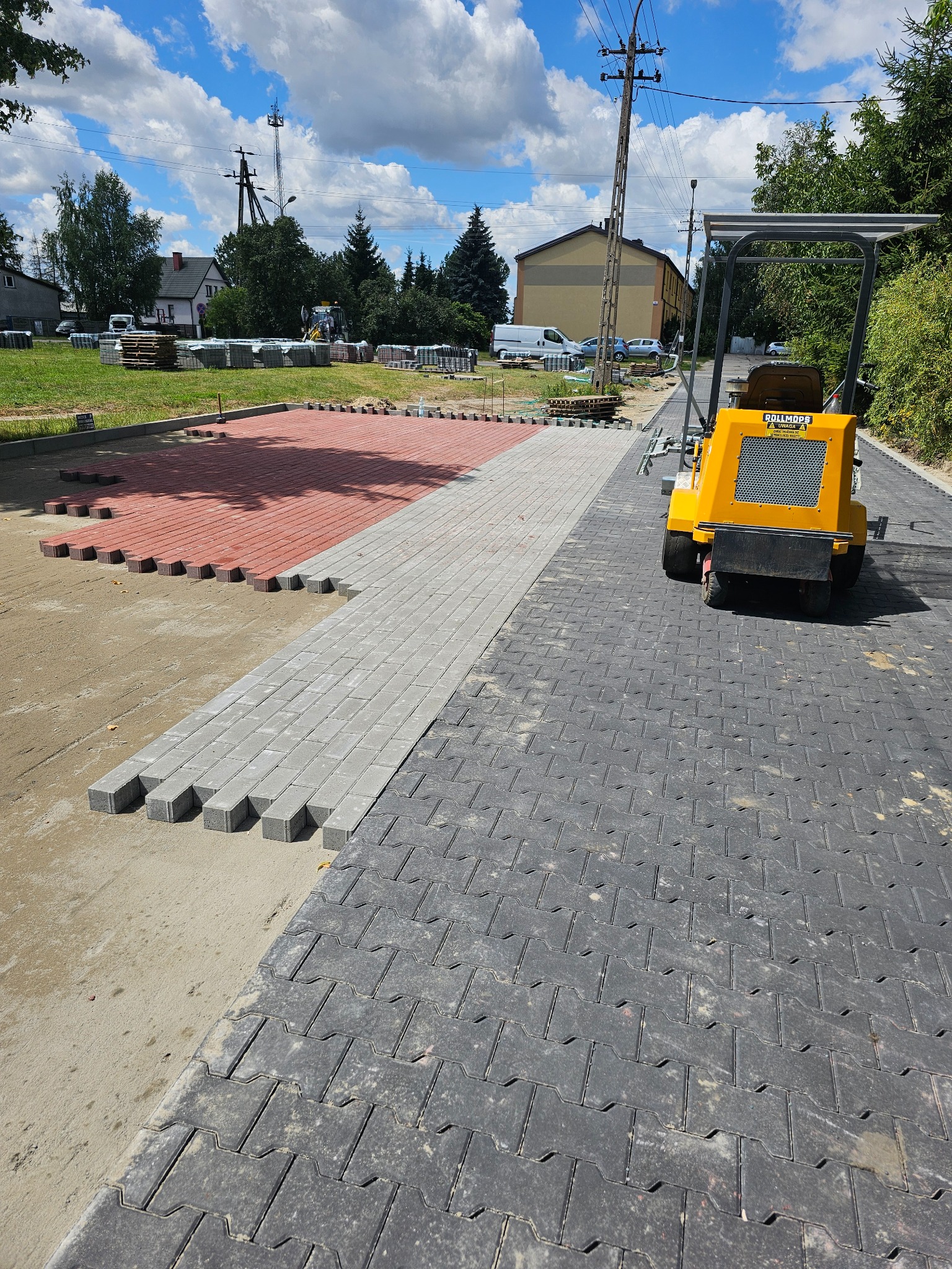 Etap prac brukarskich: częściowo ułożona, szara kostka brukowa w jodełkę, obok czerwona i szara kostka w rzędach, z tyłu maszyna do zagęszczania gruntu, w tle budynki i drzewa.