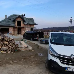 Budowa domu w stanie surowym otwartym z ułożoną dachówką, widoczne dwie osoby na dachu oraz dwa busy, jeden z przyczepką, stos drewna i desek obok budynku gospodarczego.