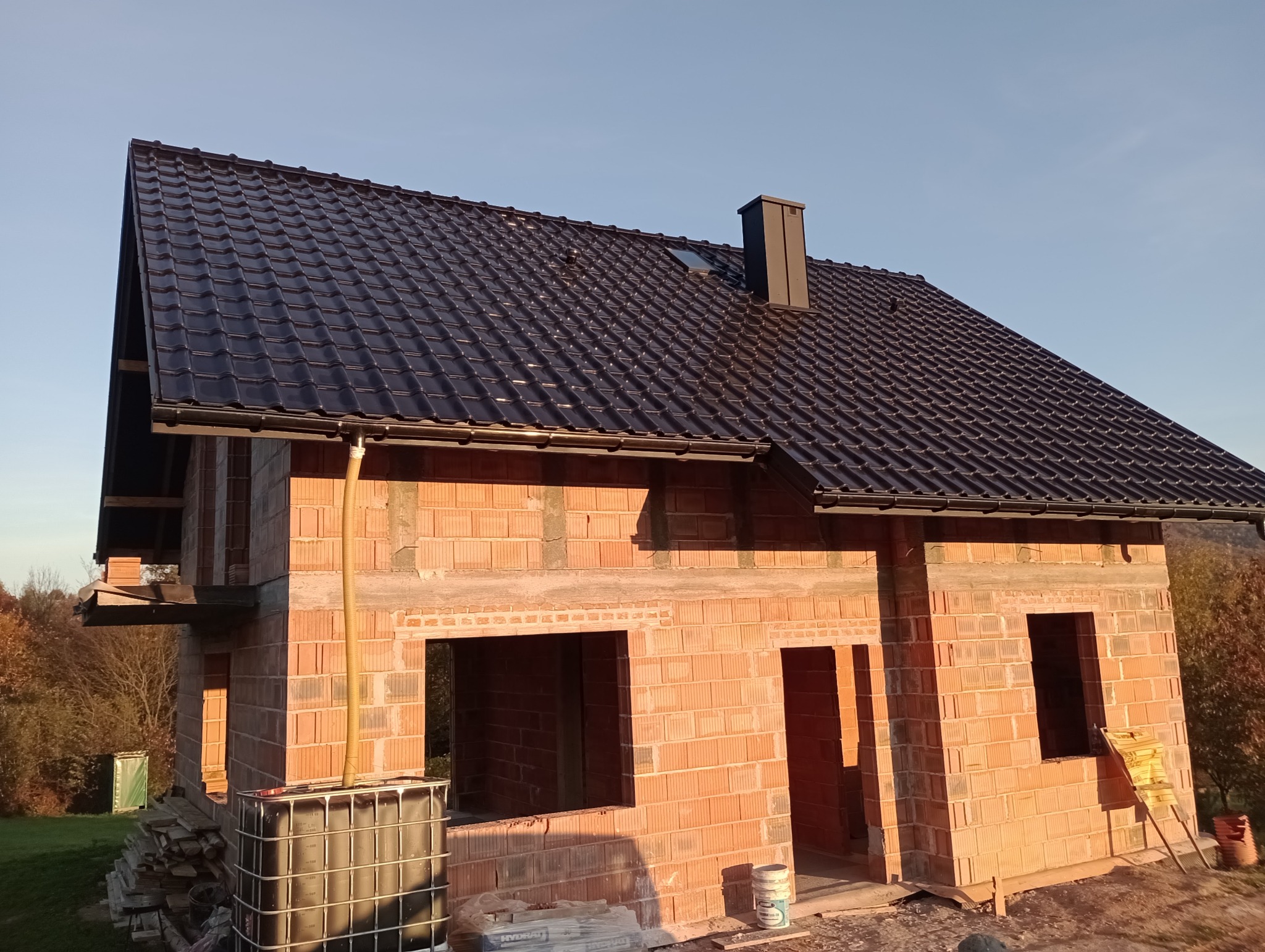 Surowy, dwukondygnacyjny dom z czerwonej cegły z zamontowaną czarną dachówką, widok z zewnątrz na tle błękitnego nieba i drzew w oddali, widoczny IBC kontener na wodę i materiały budowlane.