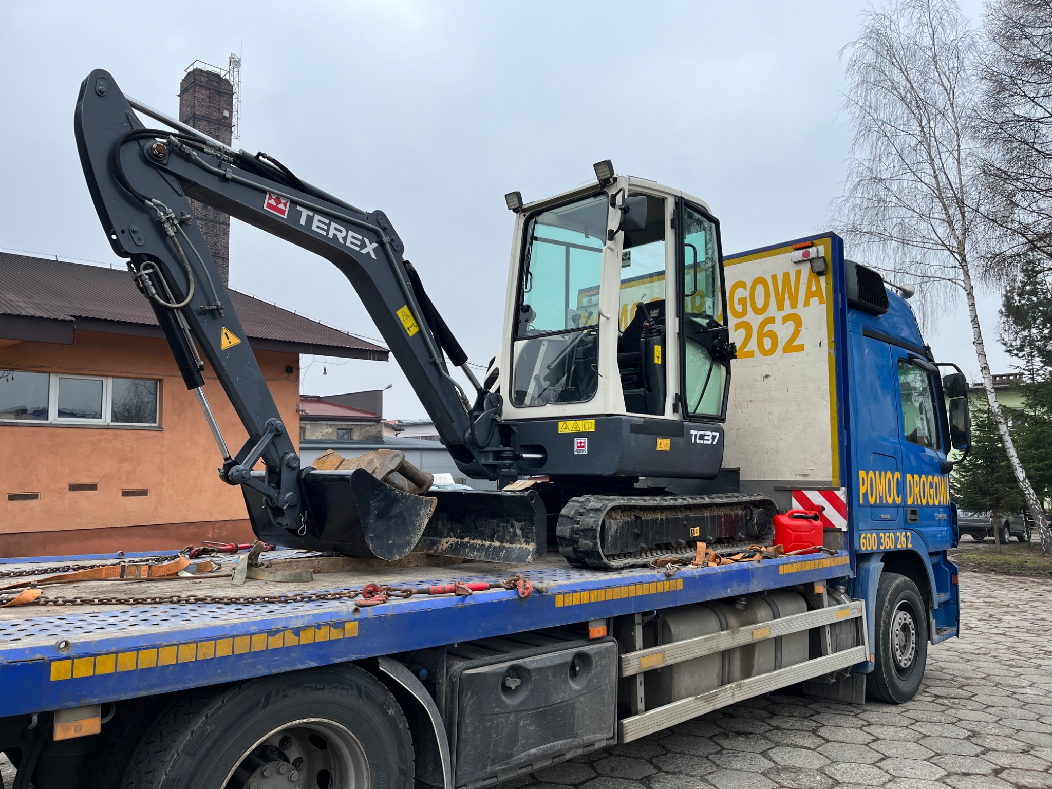 Mini koparka Terex TC37 zabezpieczona łańcuchami i pasami transportowymi na platformie lawety pomocy drogowej, widok z boku.