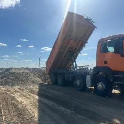 Pomarańczowa wywrotka MAN z podniesioną skrzynią ładunkową, wysypująca materiał sypki na placu budowy z turbinami wiatrowymi w tle pod błękitnym niebem.