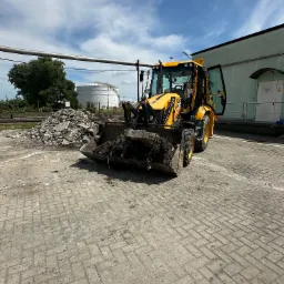 Żółta koparko-ładowarka JCB z łyżką pełną ziemi stoi na kostce brukowej, obok sterty gruzu i zbiornika. Otwarta kabina, błękitne niebo z chmurami w tle.