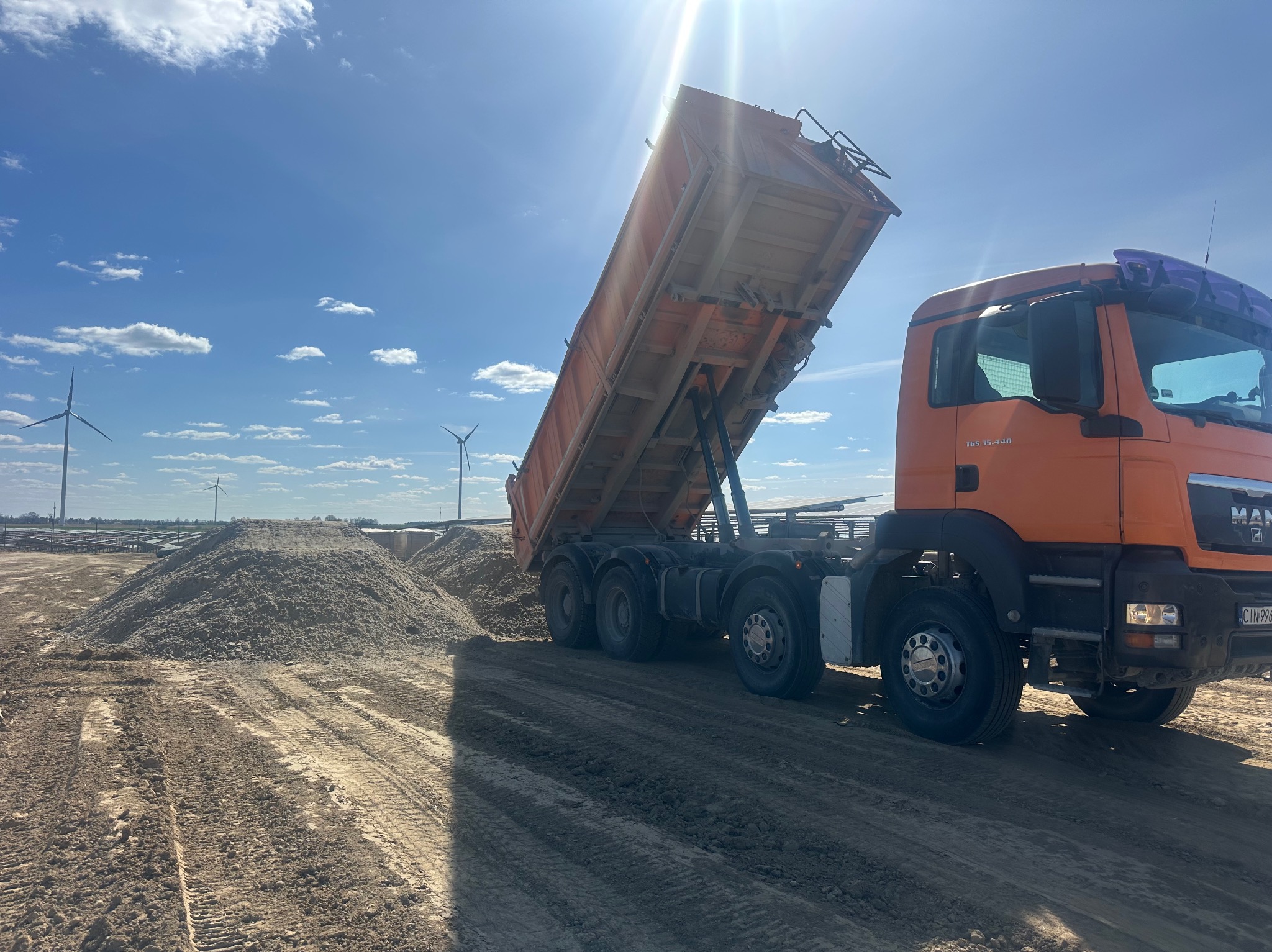 Pomarańczowa wywrotka MAN z podniesioną skrzynią ładunkową, wysypująca materiał sypki na placu budowy z turbinami wiatrowymi w tle pod błękitnym niebem.