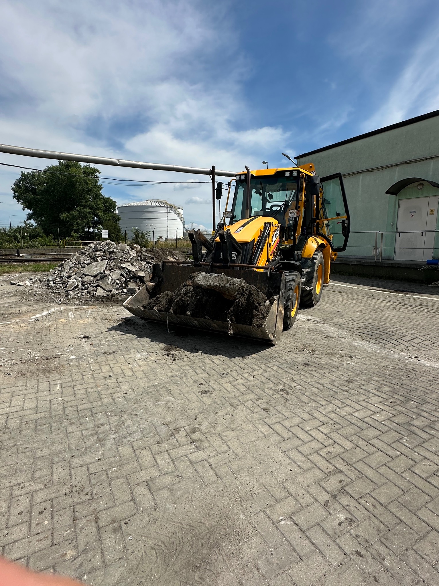 Żółta koparko-ładowarka JCB z łyżką pełną ziemi stoi na kostce brukowej, obok sterty gruzu i zbiornika. Otwarta kabina, błękitne niebo z chmurami w tle.