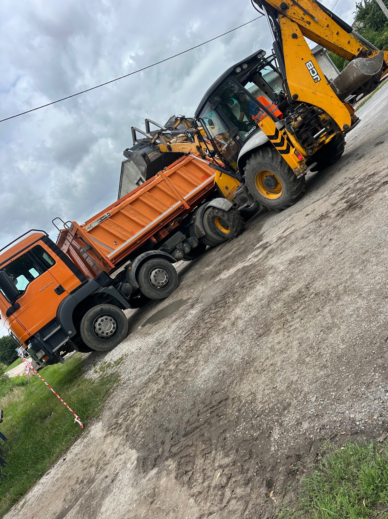 Żółta koparko-ładowarka JCB ładuje pomarańczową wywrotkę na placu budowy. Widoczne błotniste podłoże i fragment trawnika. Pochmurne niebo w tle.