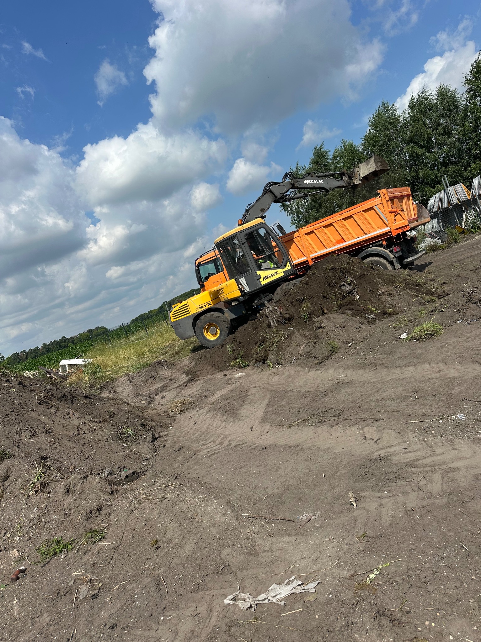 Żółto-pomarańczowa koparko-ładowarka Mecalac nabiera ziemię i wsypuje ją do pomarańczowej ciężarówki na nierównym terenie z zielonymi drzewami w tle.