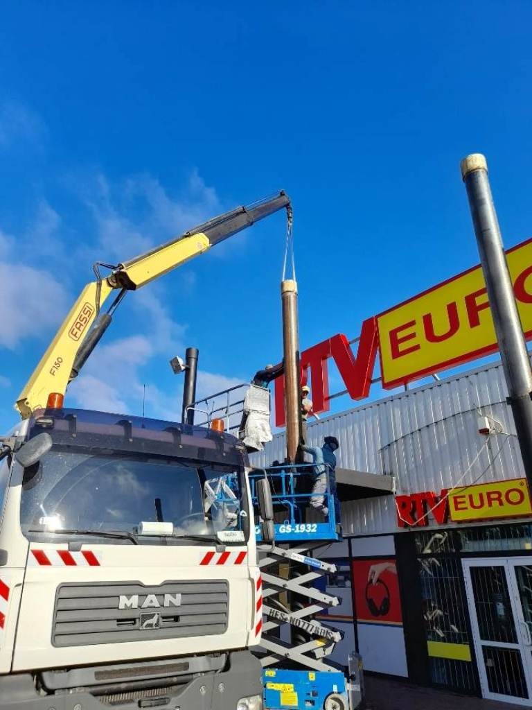 Montaż słupa konstrukcyjnego za pomocą dźwigu samochodowego i podnośnika nożycowego na tle budynku z szyldem reklamowym.