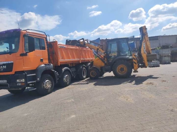 Pomarańczowa wywrotka MAN i żółto-czarna koparko-ładowarka JCB na placu budowy, przygotowane do załadunku materiału sypkiego, w tle widoczne elementy konstrukcyjne.