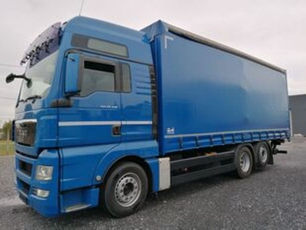 Niebieska ciężarówka MAN z naczepą kurtynową, zaparkowana na żwirowym podłożu, widok z boku na tle pochmurnego nieba.