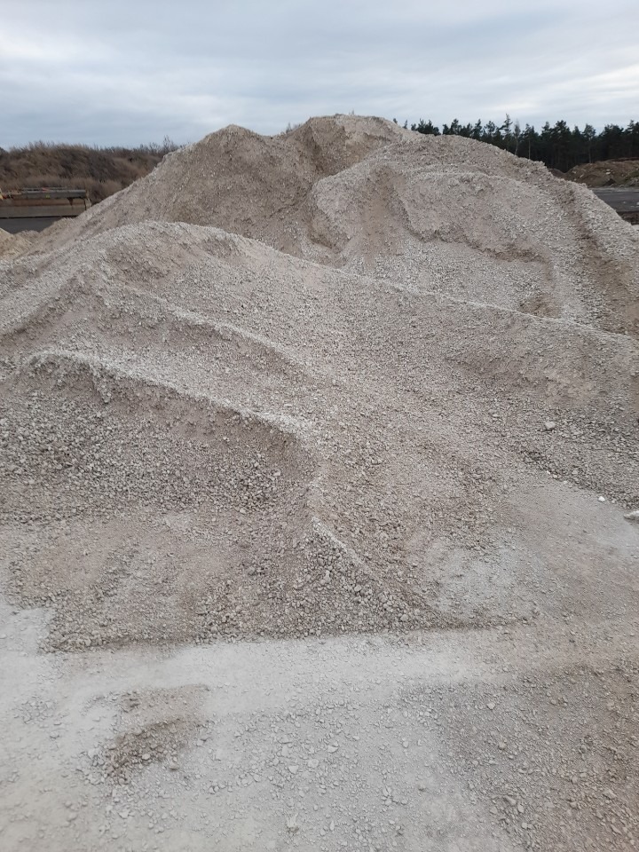 Duża pryzma szarego kruszywa o zróżnicowanej granulacji, uformowana w pagórkowaty kształt na placu składowym, z widocznymi śladami po przesypywaniu i elementami konstrukcyjnymi w tle.