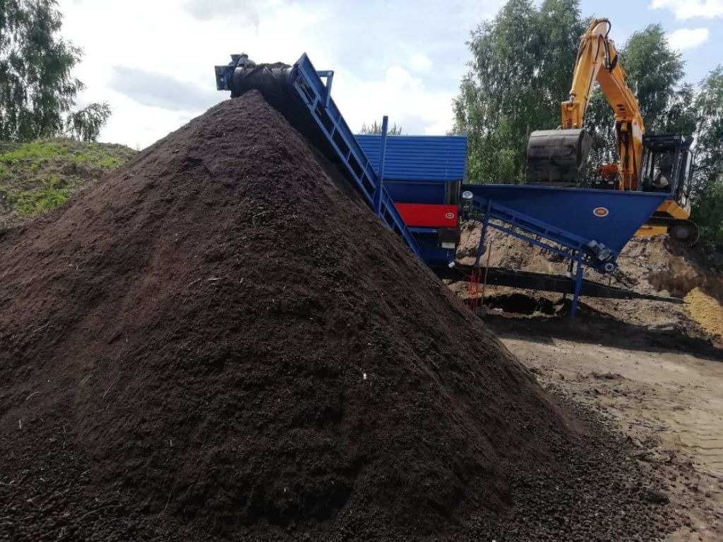 Góra ciemnej ziemi ogrodowej usypana przez podajnik taśmowy, w tle koparka i przesiewacz do ziemi.