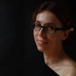 Portret uśmiechniętej kobiety w okularach, z czerwonymi i białymi kwiatami we włosach, na czarnym tle.