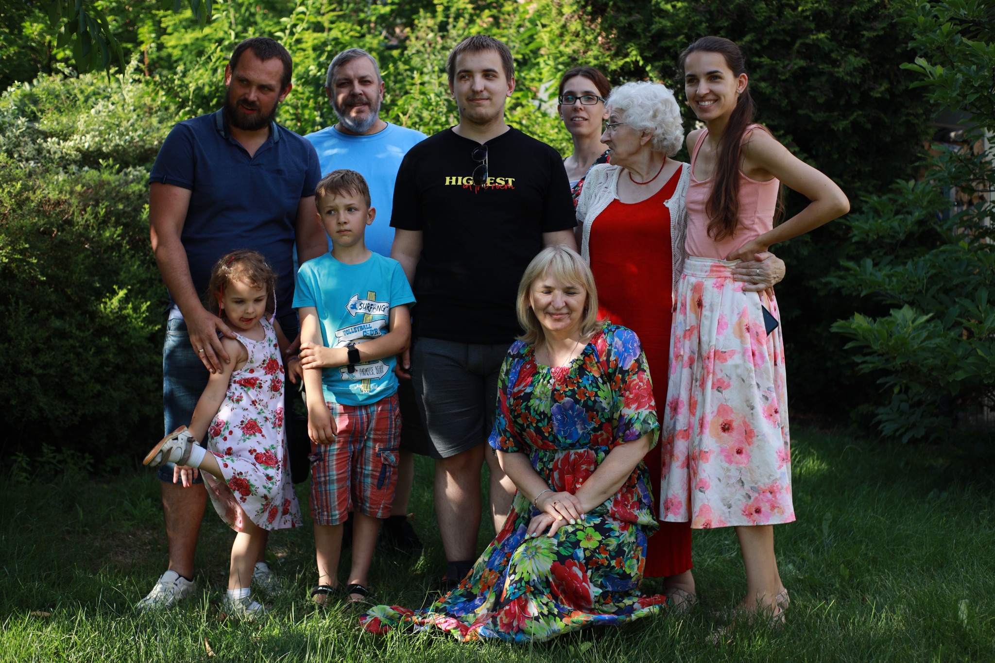 Grupa ośmiu osób w różnym wieku pozuje do zdjęcia na tle zieleni, kobieta w kwiecistej sukience klęczy na pierwszym planie.