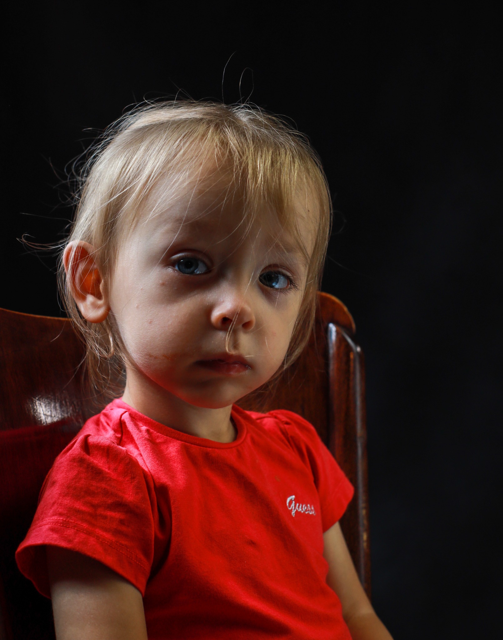 Portret skupionej, małej dziewczynki z blond włosami i niebieskimi oczami, siedzącej na drewnianym krześle na tle czarnego tła, ubrana w czerwoną koszulkę z krótkim rękawem i napisem 'Guess'.