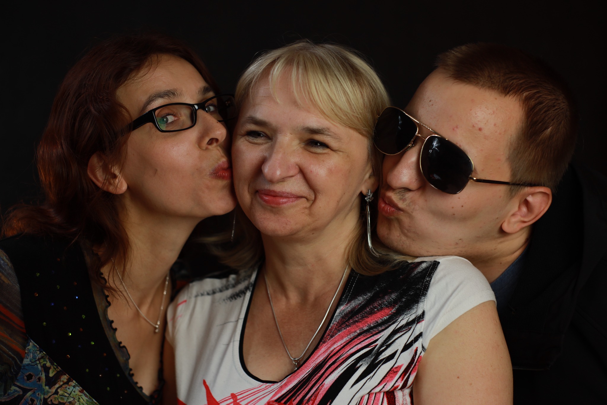 Kobieta z blond włosami uśmiecha się, całowana w policzki przez kobietę w okularach i mężczyznę w okularach przeciwsłonecznych na ciemnym tle.