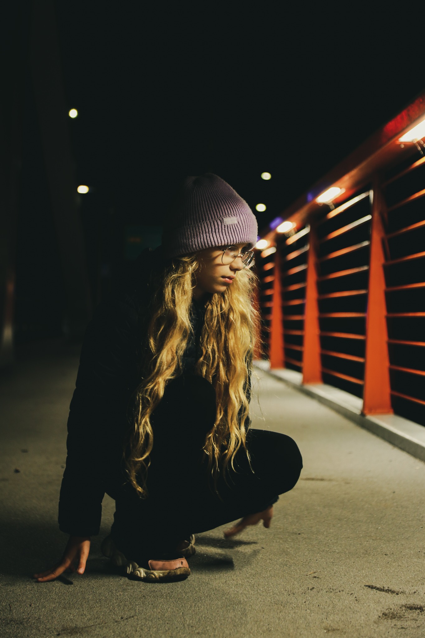 Młoda kobieta w okularach i fioletowej czapce kuca na chodniku, oświetlona światłem latarni, z pomarańczową balustradą w tle.