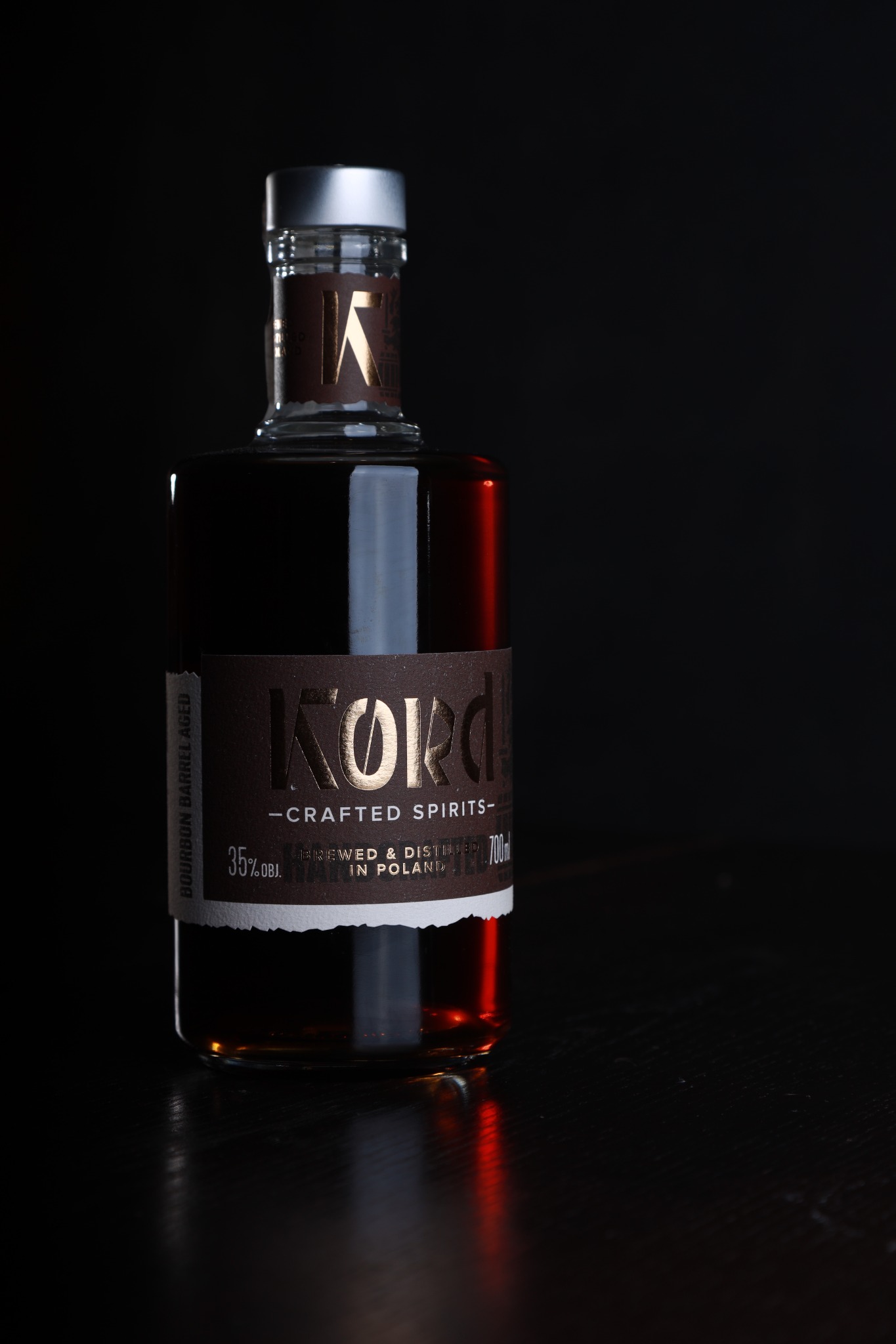 Butelka bursztynowego alkoholu Kord Crafted Spirits na ciemnym tle z refleksami światła na lakierowanym blacie.