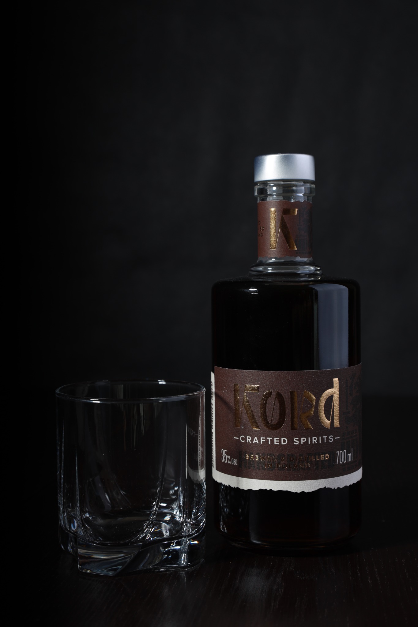Butelka ciemnego likieru Kord obok pustej szklanki z fasetowanym dnem, na czarnym tle.