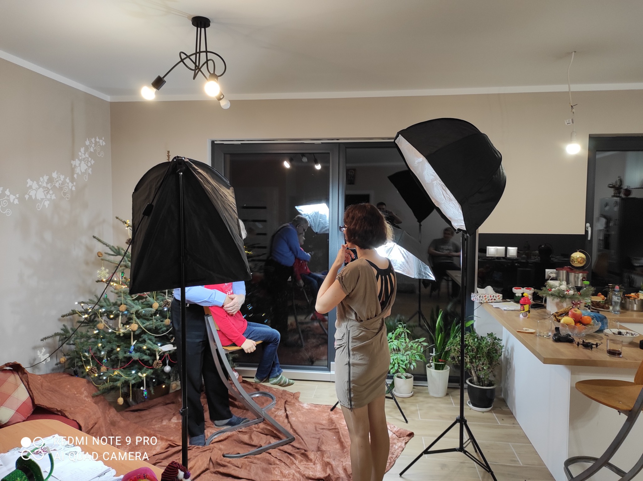 Zdjęcie zza kulis sesji w domu: oświetlenie studyjne, fotografka w beżowej sukience ustawia kadr parze siedzącej na krześle na tle choinki.