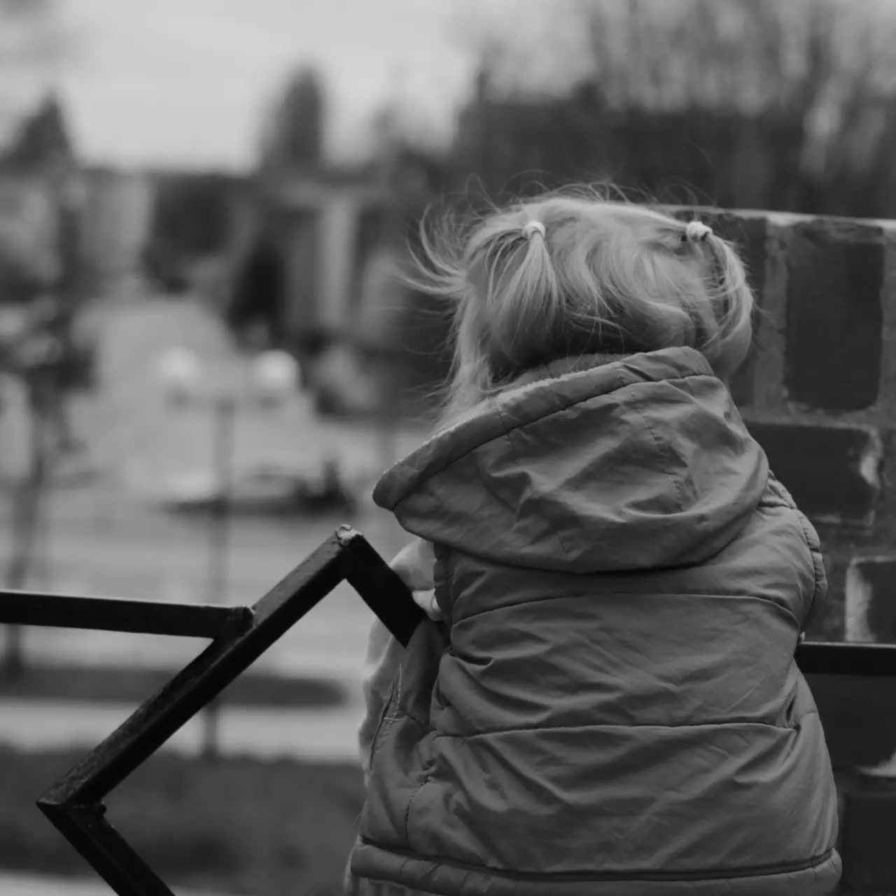 Czarno-białe zdjęcie dziewczynki w kurtce stojącej przy metalowej barierce, z warkoczykami i rozwianymi włosami, patrzącej na miasto w tle.
