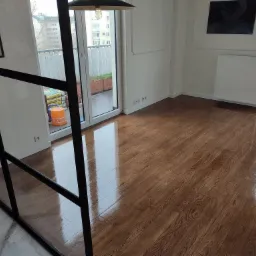 Błyszcząca, świeżo wykończona podłoga z desek w jasnym pomieszczeniu z balkonem, widoczna przez czarną, metalową ramę.