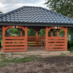 Zadaszona altana ogrodowa z ciemną dachówką, oświetleniem i drewnianą konstrukcją, obok zaparkowany skuter.