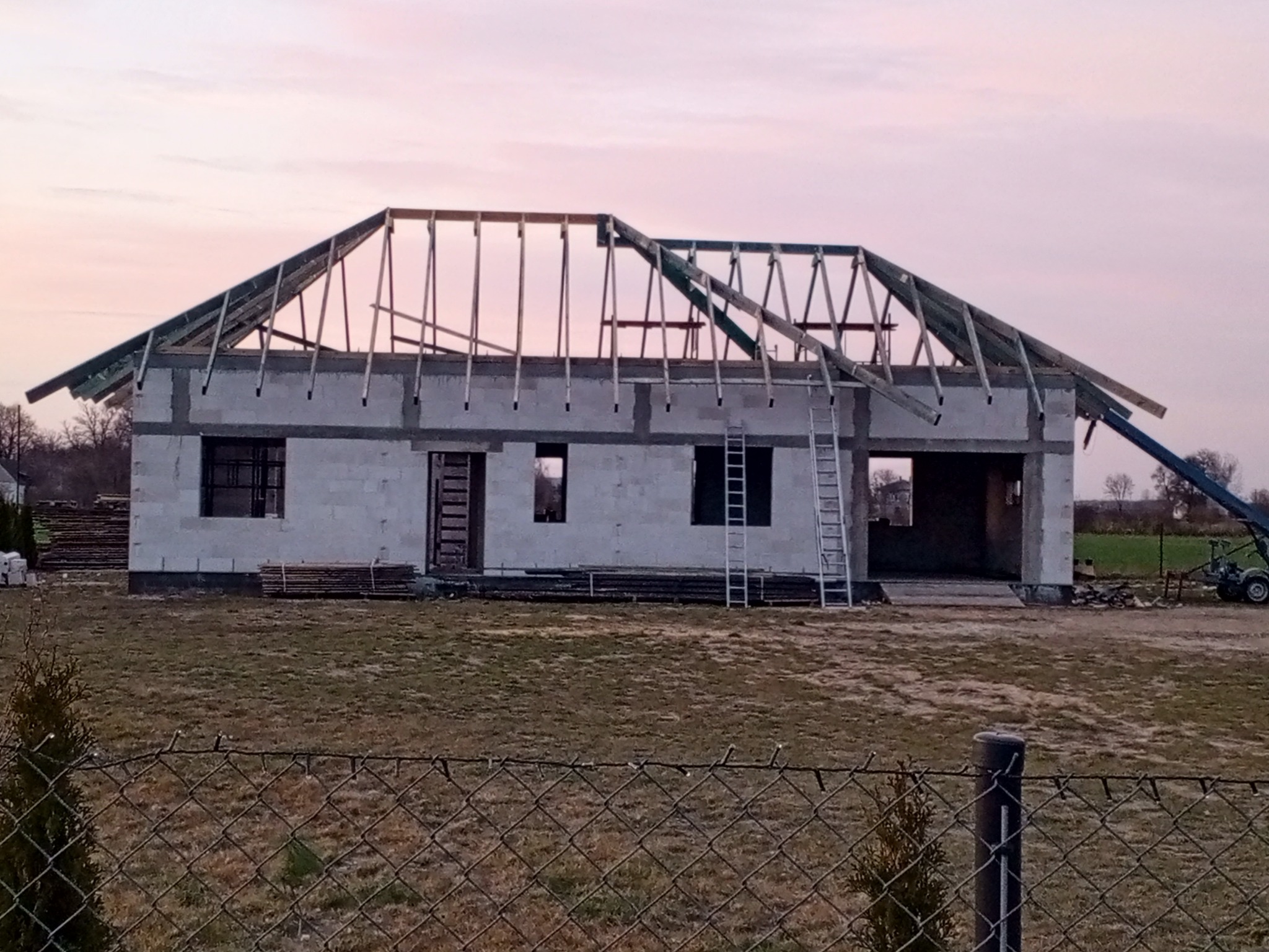 Dom w trakcie budowy, widoczna konstrukcja drewniana dachu bez pokrycia, ściany z bloczków, drabina oparta o budynek, w tle zachodzące słońce.