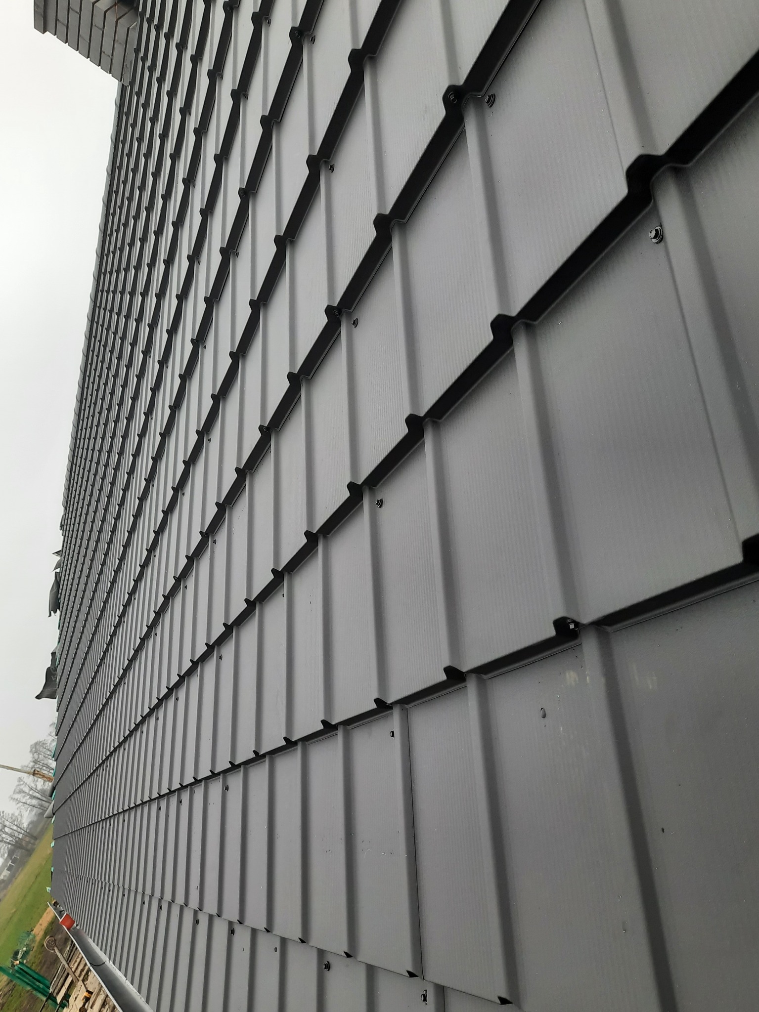 Fragment nowo położonego dachu z szarej blachy trapezowej, widok z dołu pod kątem, z widocznymi łączeniami i śrubami mocującymi, na tle pochmurnego nieba i drzew w oddali.