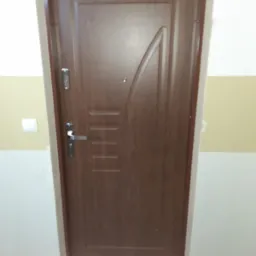 Brązowe drzwi wejściowe z metalową ościeżnicą, wyposażone w wizjer i zamek wielopunktowy, zamontowane w korytarzu z pomalowanymi na biało i żółto ścianami.