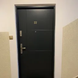 Ciemne drzwi wejściowe z numerem 22 w bloku mieszkalnym, widoczne zabezpieczenia i klamka.